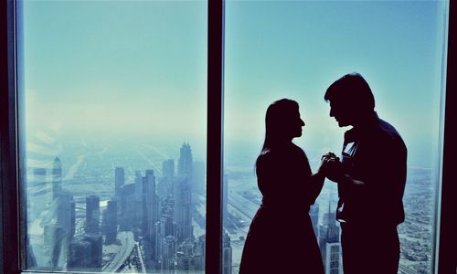 Couple holding hands while standing by glass window of building in city