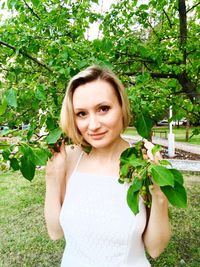 Portrait of young woman smiling while standing by tree at park