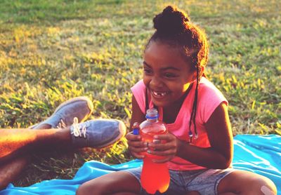 Cute girl holding cold drink bottle while sitting on picnic blanket at park