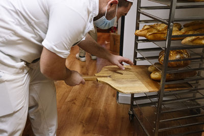 Male baker keeping bread loaf on rack with pizza peel in commercial kitchen at bakery