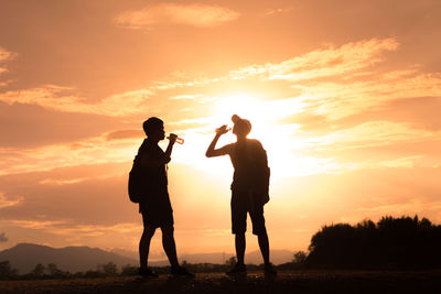 Silhouette men drinking water while standing against sky during sunset