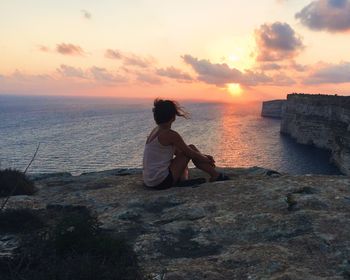 Man sitting on rock at beach against sky during sunset