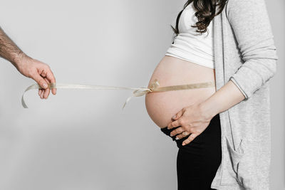 Cropped hand of man holding ribbon tied up to pregnant woman stomach against wall