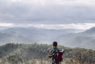 Rear view of man with dog in backpack drinking coffee while contemplating mountain landscape