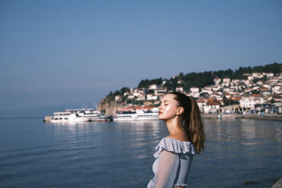 Smiling young woman standing by sea against sky in city