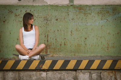 Young woman looking away while sitting on skateboard against wall