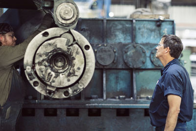 Man looking at coworker using machine at steel factory