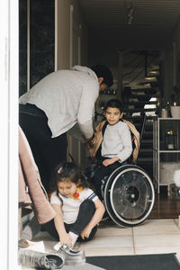 Father assisting son in wearing jacket while daughter sitting on floor at doorway