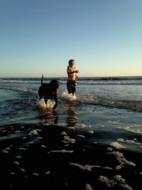 Boy with dog running at beach against clear blue sky