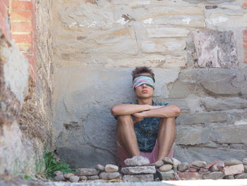 Man with covered eyes sitting against old weathered wall