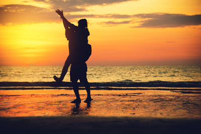 Silhouette man piggybacking woman while walking at beach during sunset