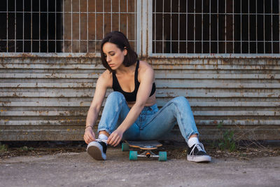 Woman tying shoelace while sitting on skateboard by fence