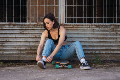 Woman tying shoelace while sitting on skateboard by fence