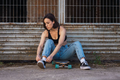 Full length of young woman sitting on skateboard tying shoelace outdoors