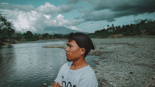 Side view of young man standing by river against cloudy sky