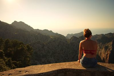 Rear view of woman sitting on retaining wall against mountains