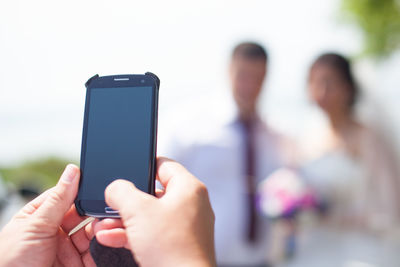 Cropped hands of person photographing people through mobile phone against sky