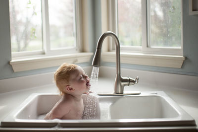 Cute baby boy sticking out tongue under running water from faucet in kitchen sink