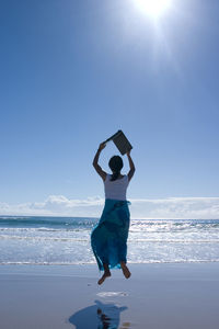 Rear view of mid adult woman holding laptop while jumping at beach against clear sky