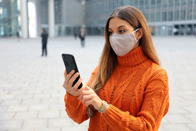 Young woman wearing mask using mobile phone while standing outdoors
