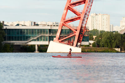 Side view of woman standing on paddle board in river and rowing with paddle under bridge in city