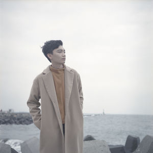 Thoughtful young man with hands in pockets looking away while standing at beach against sky