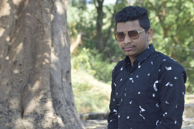 Portrait of young man wearing sunglasses standing on tree trunk
