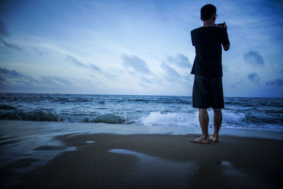 Rear view of young man photographing sea while standing at beach against sky during sunset