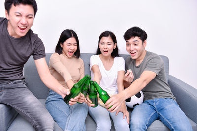 Friends toasting drinks while watching sports on sofa against wall