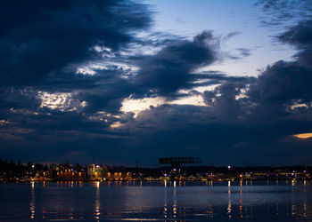 Lake by illuminated city against cloudy sky during sunset