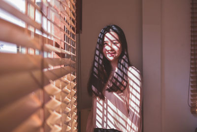 Portrait of smiling young woman with long hair standing by window blinds at home