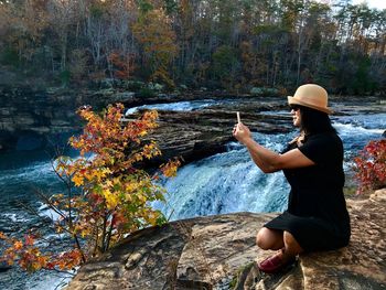 Woman photographing plants while sitting on rock by river at forest