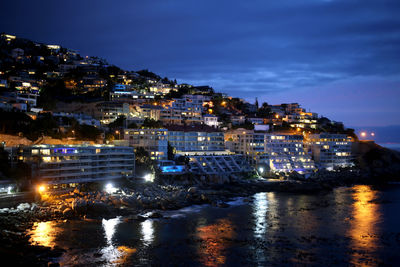 Illuminated buildings by sea against sky at night