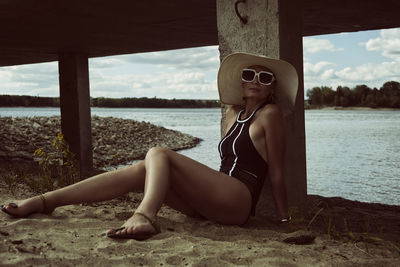 A woman in a swimsuit, hat and sunglasses sits on the sand on the riverbank at a concrete pier