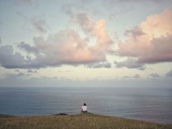 Rear view of woman sitting on mountain by sea against cloudy sky