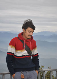 A young guy looking down, with hands in pocket while standing against the background of mountains