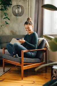 Mid adult woman smiling while using mobile phone on armchair at home