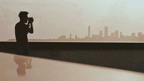 Silhouette man photographing against sky in city during sunset