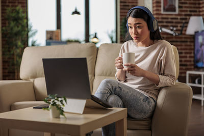 Businesswoman wearing wireless headphones looking at laptop sitting on sofa