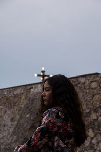 Side view of thoughtful young woman looking away while standing by wall against sky