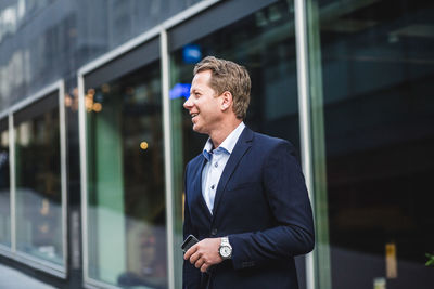 Smiling mature businessman with smart phone looking away while standing against office building