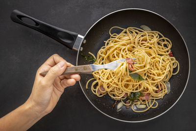 Hand holding fork get spaghetti pasta with dried chilli, garlic, sweet basil and bacon in black pan