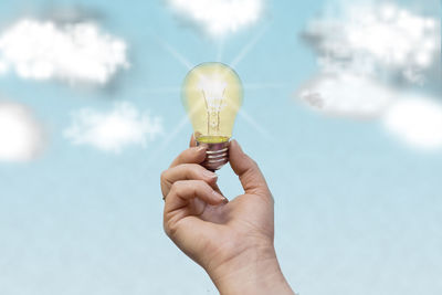 Close-up of hand holding light bulb against sky