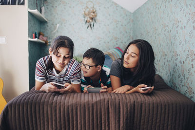 Siblings using wireless technologies while lying on bed by wall at home