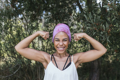 Happy woman with purple bandana flexing muscles while standing against plants at park