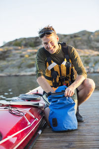 Portrait of happy man searching something in bag by kayak on pier