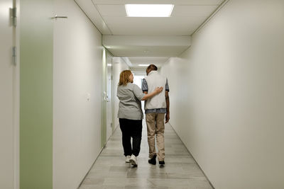 Full length rear view of female nurse walking with arm around senior man in corridor at retirement home