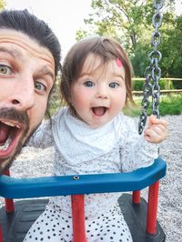 Close-up portrait of shocked man with cute toddler daughter on swing at playground