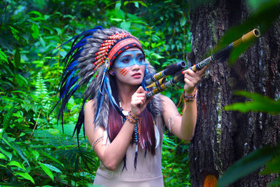 Young woman with face paint aiming weapon while standing in forest