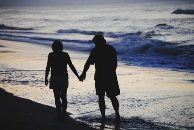 Rear view of silhouette couple holding hands while walking at beach during sunset